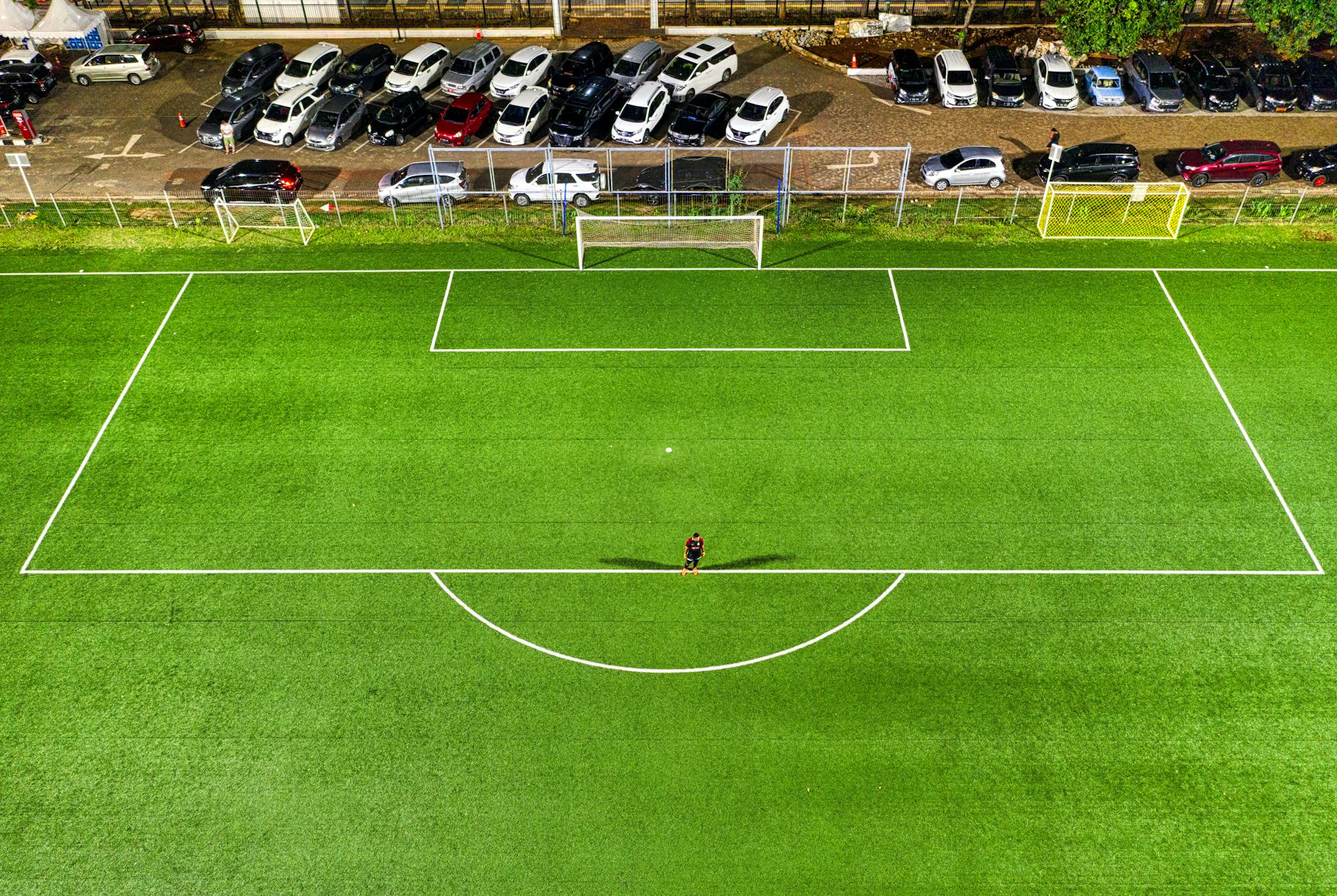Aerial view of a soccer field at night in Jakarta with a lone player casting a shadow under stadium lights.