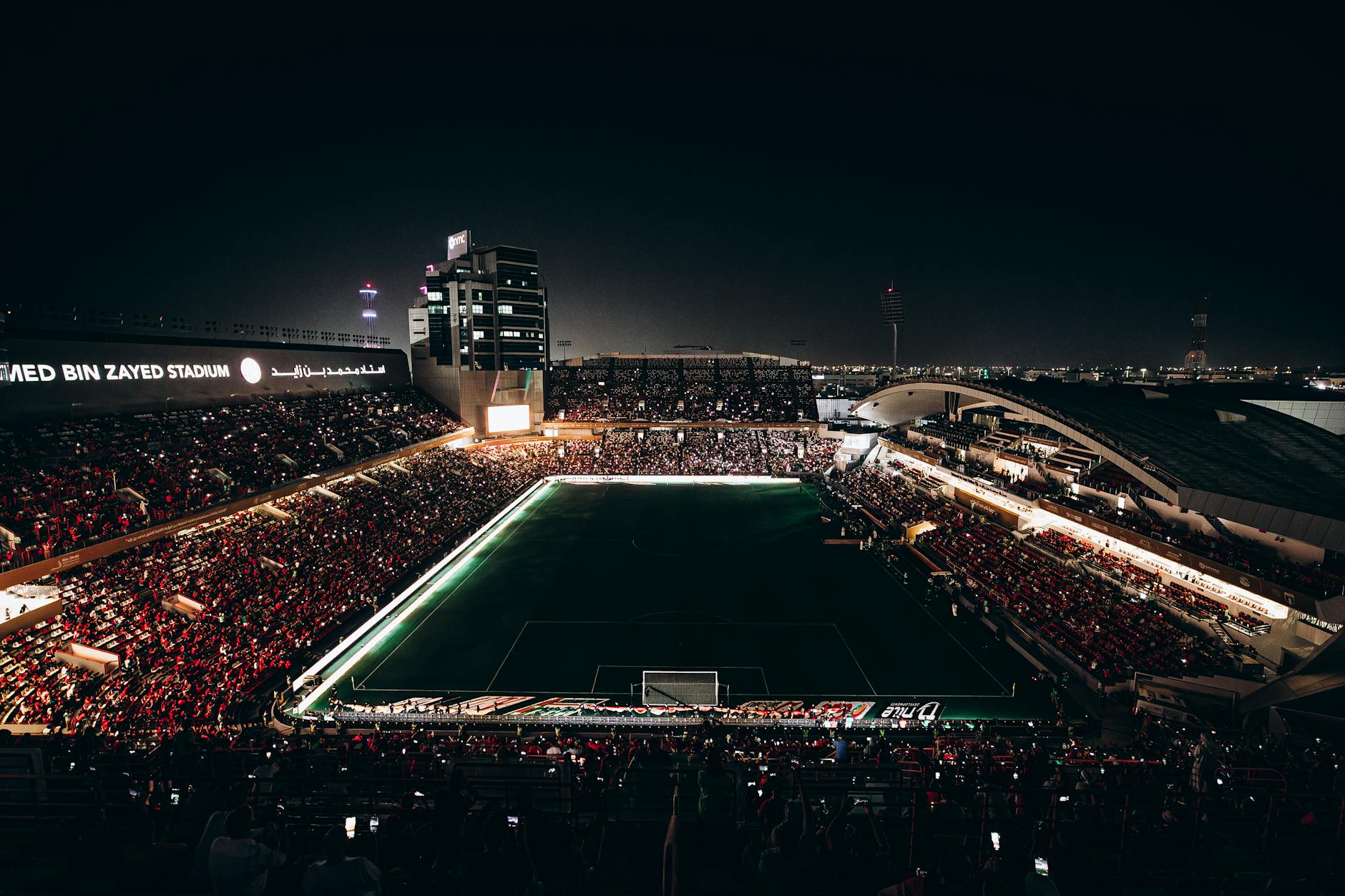 A vibrant nighttime aerial view of a football match at Mohammed Bin Zayed Stadium in Abu Dhabi.