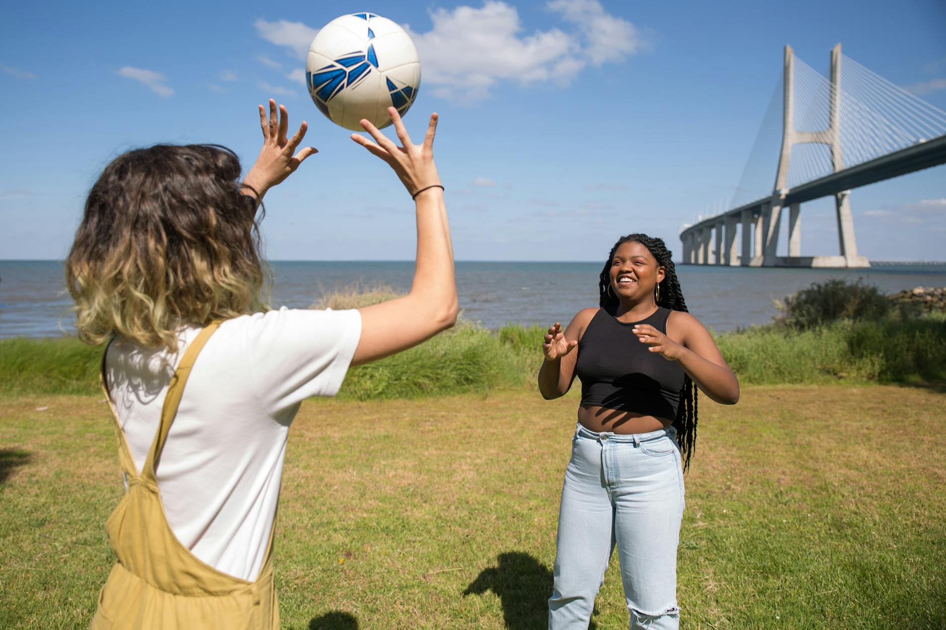 Two women enjoying a game of volleyball by Vasco da Gama Bridge under clear skies.
